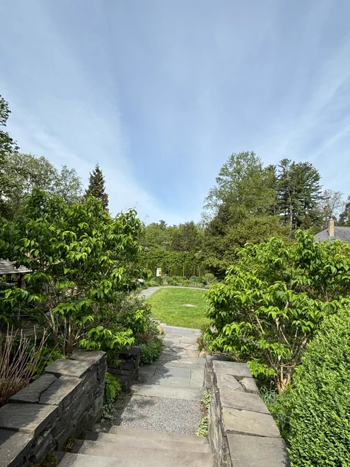 Stone steps bordered by low stone walls lead into a lush garden with manicured shrubs, a curved pathway, and a circular lawn under a bright blue sky.