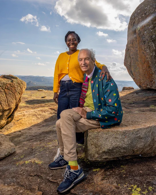 Jon Snow, an older white man wears a colourful jacket and tie, and has tan trousers. Precious, a Black woman, wears a yellow cardigan and jeans. She is standing behind him with her arm on his shoulder as he sits on a large rock. They are outdoors on a rocky landscape with distant mountains under a cloudy sky. 