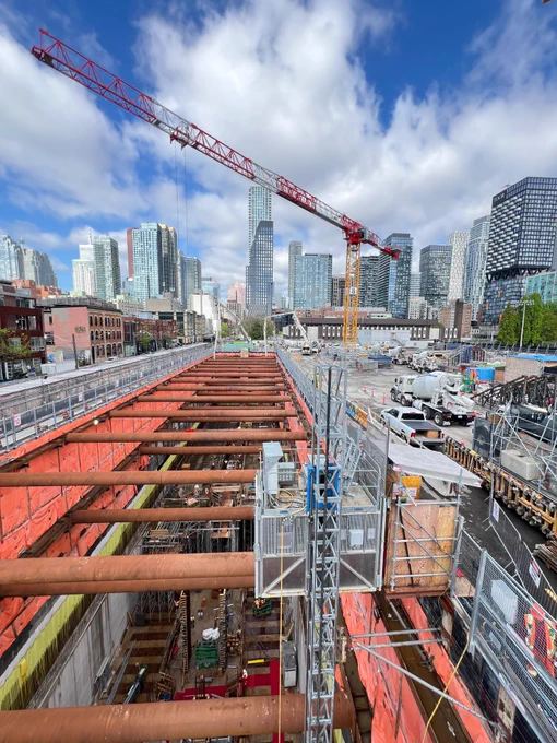 A busy construction site in an urban area with a red construction crane and several tall city buildings in the background. The site is bordered by orange barriers and contains scaffolding and large pipes. The sky is partly cloudy.