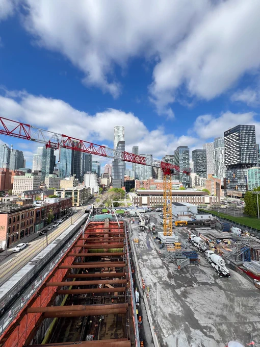 A construction site in Toronto with a large red crane and yellow crane. Skyscrapers in the background under a partly cloudy sky. 