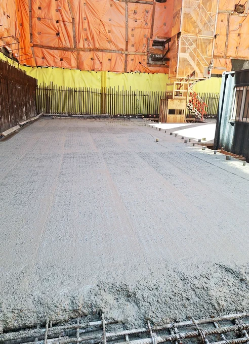 Un chantier de construction avec du béton frais coulé sur le sol. La zone est bordée de barrières orange et jaunes temporaires et d’une structure métallique. L’échafaudage et une structure recouverte de bâches de protection sont visibles à l’arrière-plan.