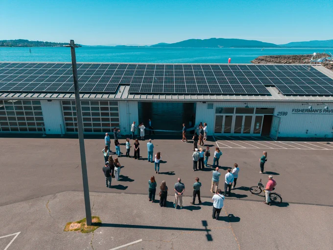 A crowd attends the ribbon cutting at the Port of Bellingham solar site while the building, it's panels, and Bellingham Bay rise into the background.