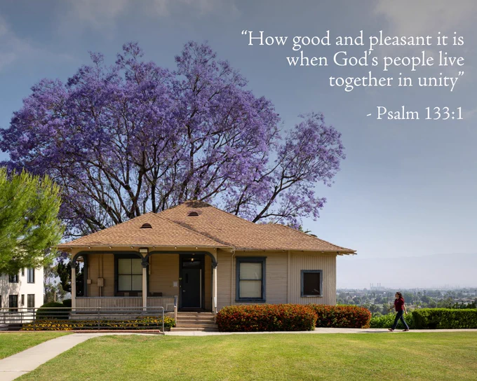 A small, single-story house with a porch sits under a large Jacaranda tree with purple blooms. A woman walks along a path in front of the house.  The background shows a cityscape under a clear sky. A quote from Psalm 133:1 is in the upper right corner, it states "How good and pleasant it is when God’s people live together in unity" 