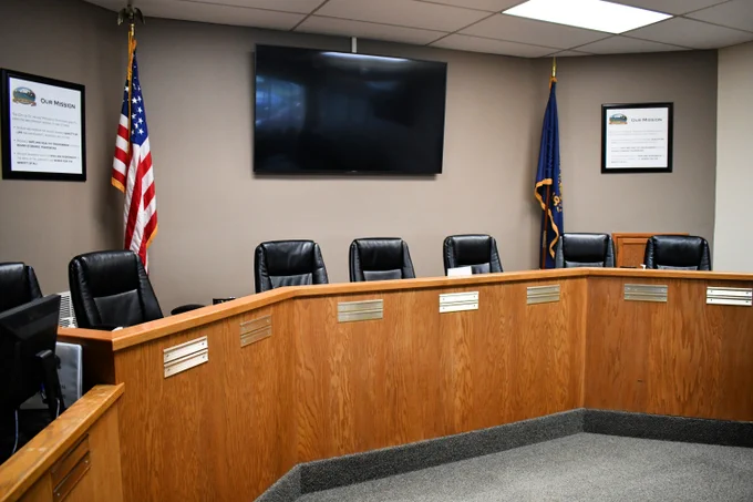 Wooden curved dais with black chairs arranged behind it and a wall mounted with a large screen tv and flanked by the United States flag and the State of Oregon flag