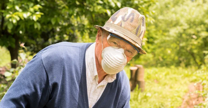An elderly individual wearing a straw hat and a dust mask works in a lush garden.