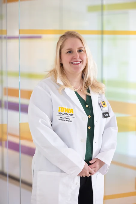A woman with long blonde hair smiles while standing in a brightly lit room. She is wearing a white lab coat with the "Iowa Health Care" and "Stead Family Children's Hospital" logos visible. Underneath, she has a green top. The background features a glass wall with horizontal stripes in purple, orange, and green.