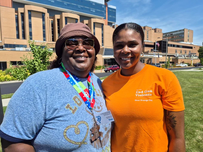 Two people are smiling outdoors in front of Sentara Norfolk General Hospital. One is wearing a brown hat and a shirt with a colorful lanyard, while the other is in an orange shirt with the message "End Gun Violence" and "Wear Orange Day.