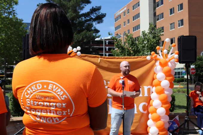A person wearing an orange T-shirt with "CHKD - EVMS - SNGH - We are Stronger Together!" written on the back stands in the foreground. Another person is speaking in front of an orange backdrop decorated with white and orange balloons. The event takes place outdoors with trees and a brick building in the background.