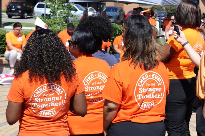 A group of people wearing bright orange T-shirts that read "CHLD EVMS SNGH We are Stronger Together!" are gathered in an outdoor setting. Some individuals are standing, while others are seated in the background. 