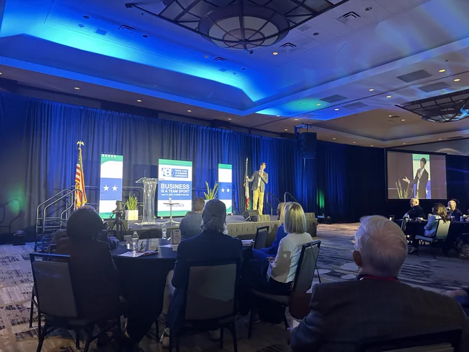 A photo of Rob speaking on stage at the ABI conference. It's being held in a large, well-lit room. The backdrop features signage with the U.S. flag and displays reading "Business in America." Attendees are seated at round tables facing the stage, and there's a projection screen displaying the speaker on the right.