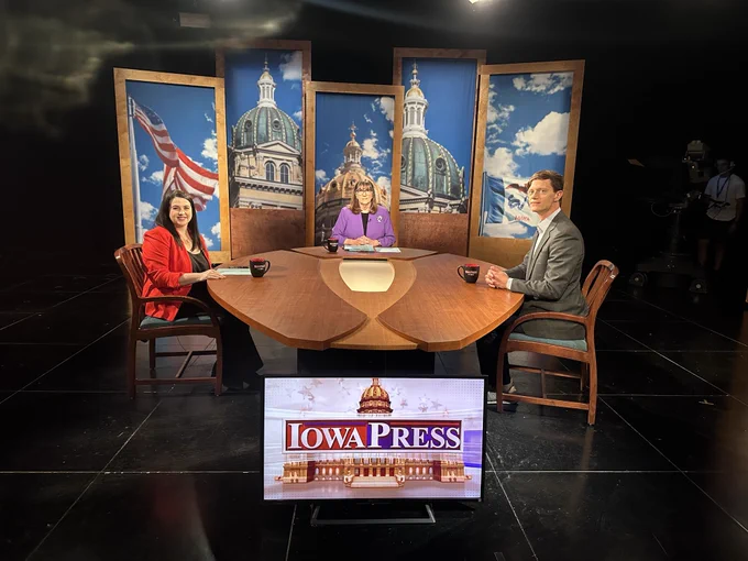 Rob and two other individuals are seated at a round wooden table in the Iowa Press studio. Behind them are three large vertical panels depicting the Iowa State Capitol and a U.S. flag. A screen in front of the table displays the "Iowa Press" logo. Camera equipment and a crew member are visible in the background.
