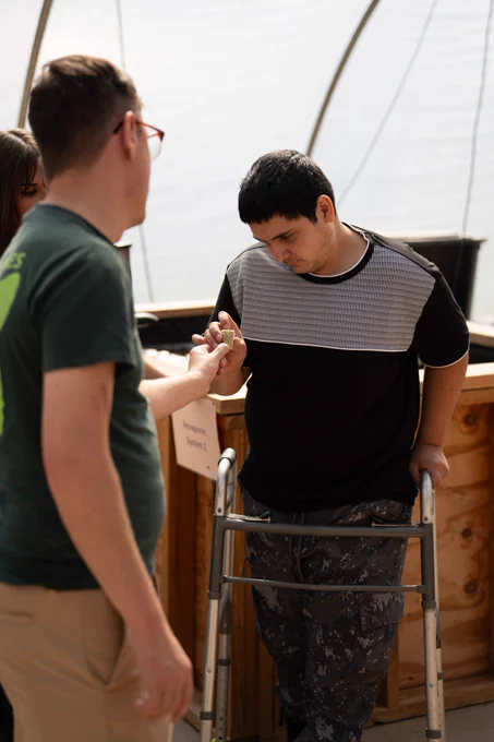 Able SC staff with disabled youth look at an item in a greenhouse, a white man with glasses in an Equip shirt, and a young man with brown skin using a walker.
