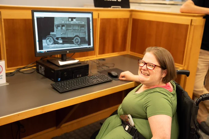 Disabled youth works at an accessible computer desk at the library, a young white woman in a wheelchair.