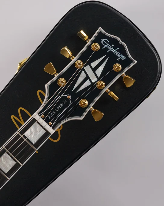 An Epiphone guitar's headstock resting on a black guitar case. The Inspired by Gibson Custom headstock features gold tuning pegs, and a signature on the guitar case reads "Alex Lifeson.