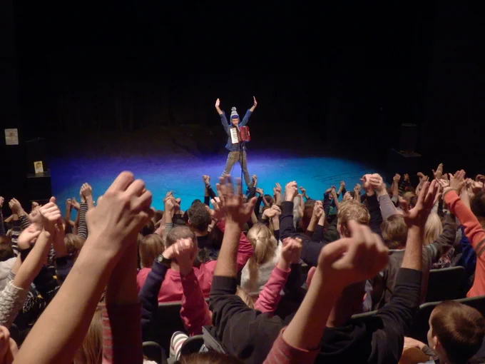 A performer on stage plays an accordion while an audience raises their hands enthusiastically in a theater setting.
