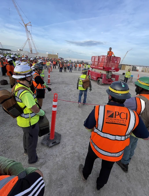Construction workers wearing safety gear gather outdoors for a safety briefing at a large job site with cranes in the background.
