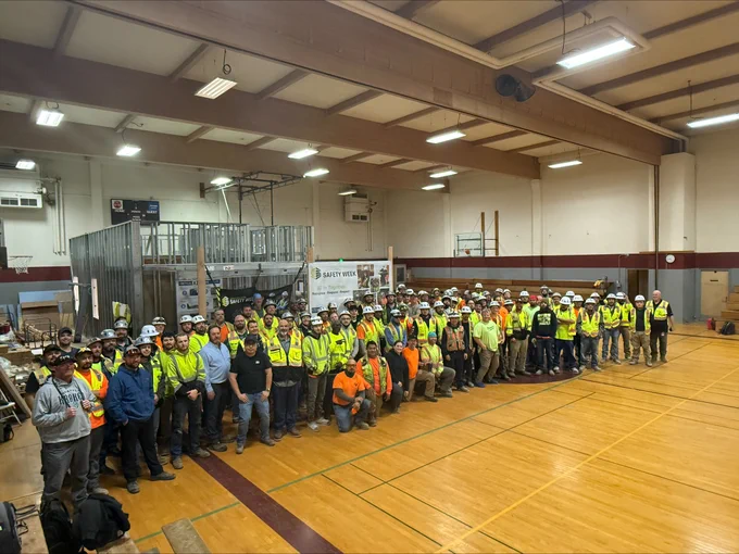 Large group of construction workers and supervisors wearing safety vests and helmets gathered inside a gymnasium for a team photo.