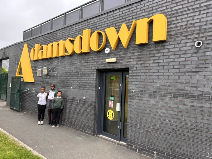 Three children stand outside Adamsdown school, one holding a certificate.