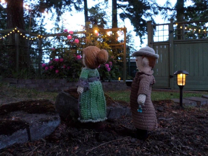 Crochet dolls of Marshal Kahler, holding earrings, standing behind Mary Woodhull, who is looking at the new solar lights, which have come on at twilight. Yard & fence in background. 