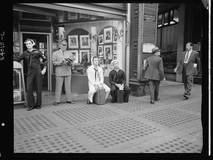 A small group of people, including two sailors sitting on suitcases, watch an electronic sign in Times Square for news about the D-Day invasion.