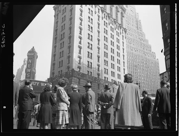 A large group of people watch an electronic sign in Times Square for news about the D-Day invasion. The photographer is behind the group, the news ticker is in view.