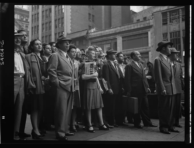 A large group of people watch an electronic sign in Times Square for news about the D-Day invasion. The photographer is between the group and the news ticker. 