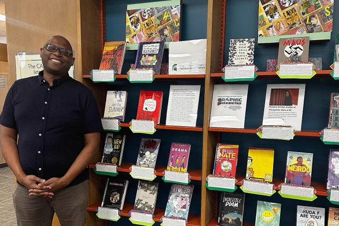 A person smiling stands beside a library book display featuring various graphic novels. The shelves are filled with colorful covers, creating a vibrant and welcoming atmosphere.