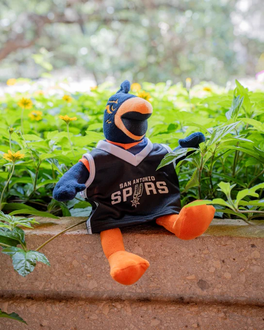 A plush toy of a blue and orange bird wearing a San Antonio Spurs jersey is sitting on a stone ledge surrounded by greenery and yellow flowers.