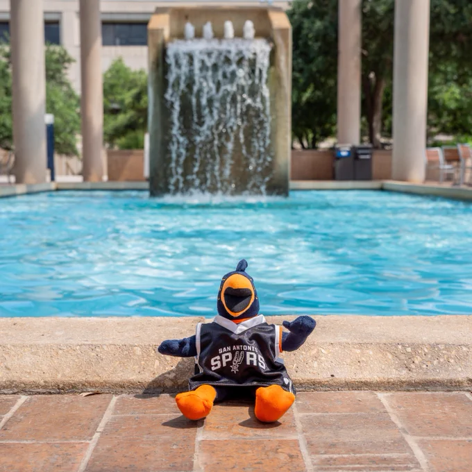 A plush toy wearing a San Antonio Spurs jersey is sitting on the edge of a fountain, with water cascading down a large stone structure in the background, surrounded by tall columns.