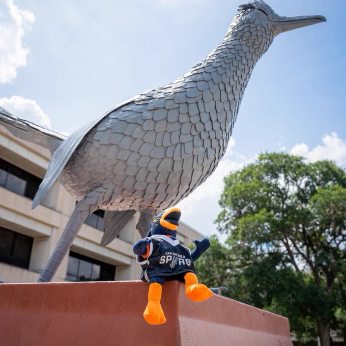 A statue of a large roadrunner stands on a pedestal. In the foreground, a toy wearing a "Spurs" jersey sits in front of the statue. Trees and a building are visible in the background under a partly cloudy sky.