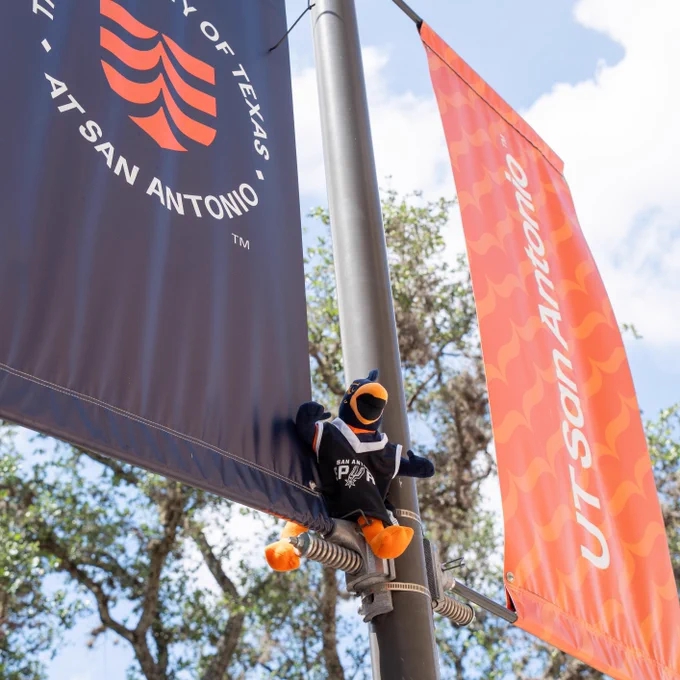 A plush bird toy wearing a dark shirt is attached to a pole with banners for The University of Texas at San Antonio. Trees and a blue sky are in the background.