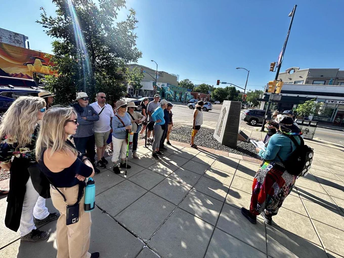 Group of people attentively listening to a guide speaking on a sunny urban sidewalk near a traffic light intersection.