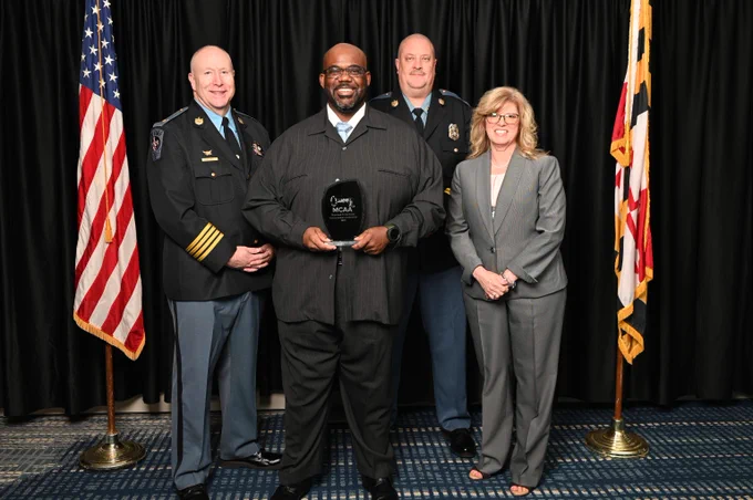 Group photo of three uniformed officers and a woman in a suit standing with an award plaque between two flags.