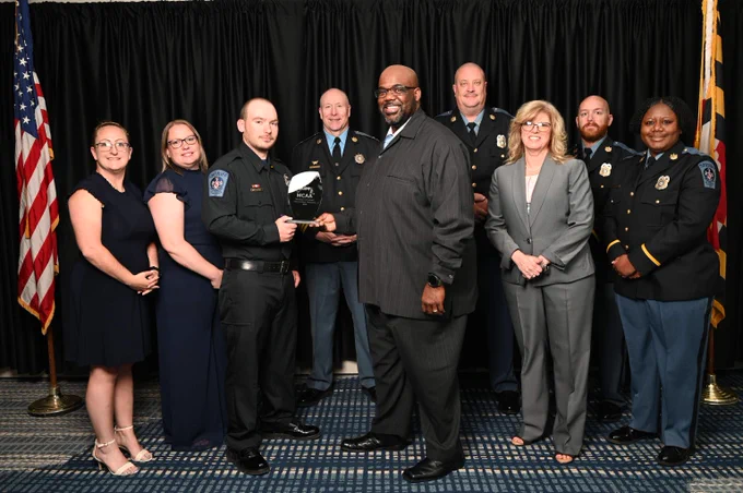 Group photo of uniformed police officers and formally dressed individuals standing together in front of black curtains with American flags on each side.