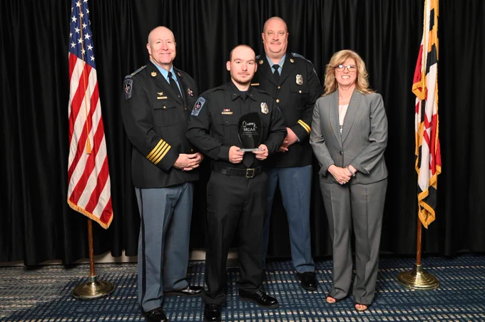 Four individuals stand in front of black curtains with American and Maryland flags on either side, three in police uniforms and one in a gray suit, holding an award plaque.
