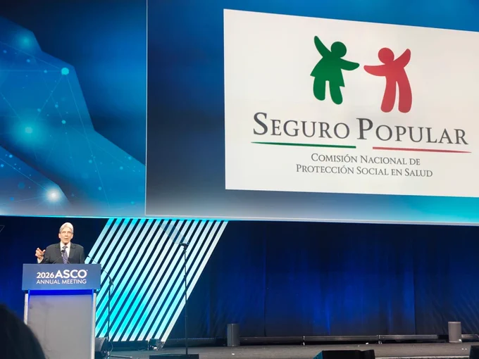 At a stage presentation at the 2026 ASCO Annual Meeting, a person speaks at a podium with ASCO branding. The large screen behind the speaker  displays the logo of "Seguro Popular," accompanied by the text "Comisión Nacional de Protección Social en Salud." The background features a modern design with blue lighting and abstract patterns.
