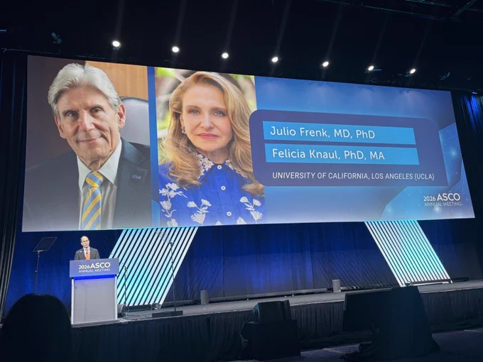 A large presentation screen at the 2026 ASCO Annual Meeting features two headshots with names and credentials: Julio Frenk, MD, PhD, and Felicia Knaul, PhD, MA, from the University of California, Los Angeles. A speaker stands at the podium below the screen in a dark, formal conference setting.