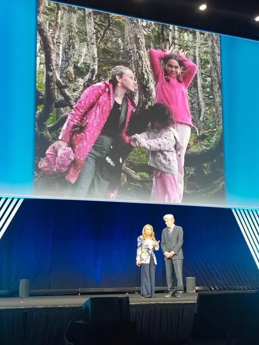 Two individuals standing on a conference stage speak in front of a large screen displaying a photograph of three people outdoors in a forested area. In the photo on the screen, one person is wearing a pink jacket, another is in a pink shirt, and a child is visible interacting with them. The stage features a formal conference setup with blue lighting and decorative elements.