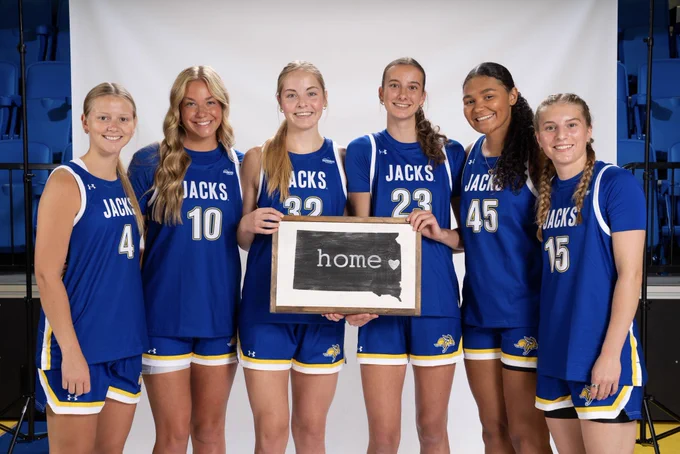 Six basketball players standing side-by-side in a gym setting, in front of a white studio photo backdrop. All are dressed in blue Jacks uniforms. The two in the center hold a sign with the outline of the state of South Dakota.