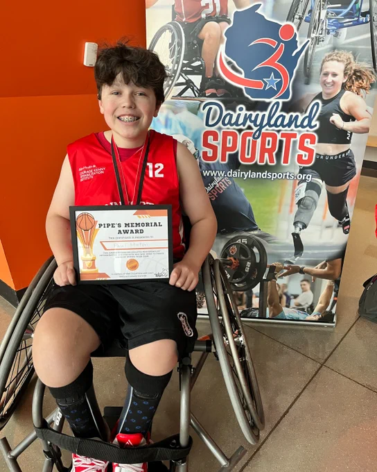 A smiling child in a wheelchair holds a "Pipe's Memorial Award" certificate, wearing a red sports jersey, in front of a Dairyland Sports banner.