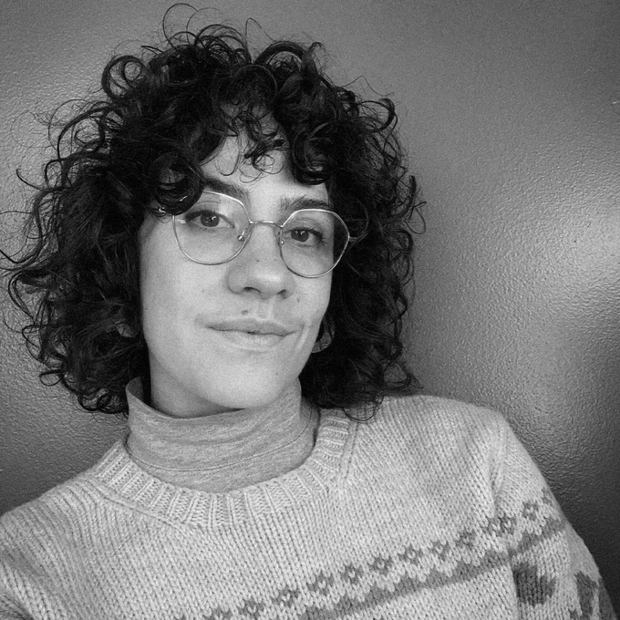 A black and white portrait of a person with curly dark hair and round glasses, wearing a turtleneck under a knit sweater, smiling faintly against a plain background.