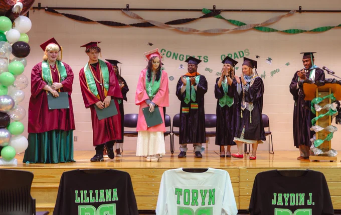 A small group of high school graduates in caps and gowns stands onstage together.
