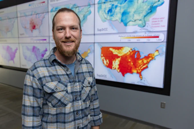 A person stands smiling in front of a wall displaying colorful climate maps of the United States. 