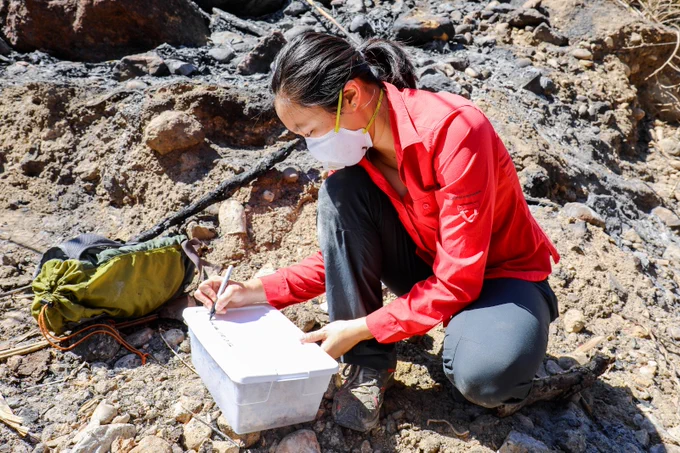 A person wearing a red shirt and a face mask is crouched on rocky terrain, writing on a clipboard. A green backpack rests nearby.