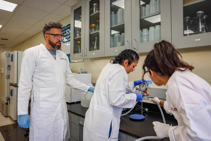 Three individuals in lab coats work together in a laboratory setting. One holds equipment, while another observes. Shelves with bottles and equipment are visible in the background.
