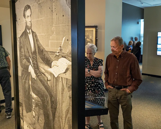 Visitors examine a museum display case behind a large illustration of Abraham Lincoln.