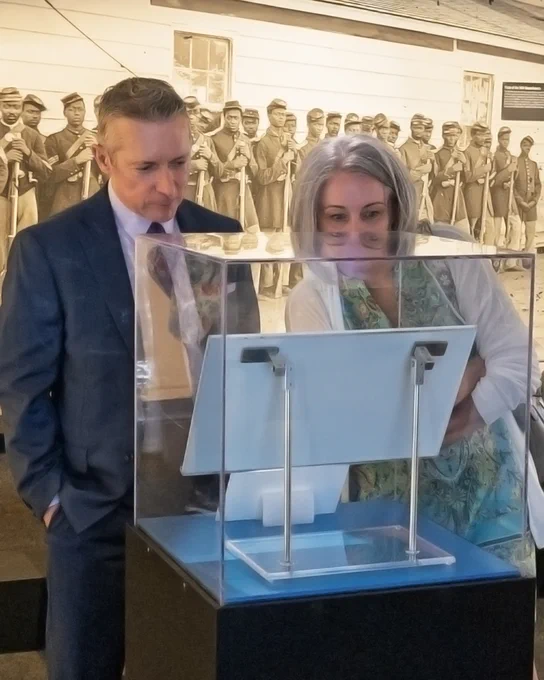 Two visitors closely examine a historical exhibit encased in glass at a museum with a large black-and-white photo of soldiers in the background.