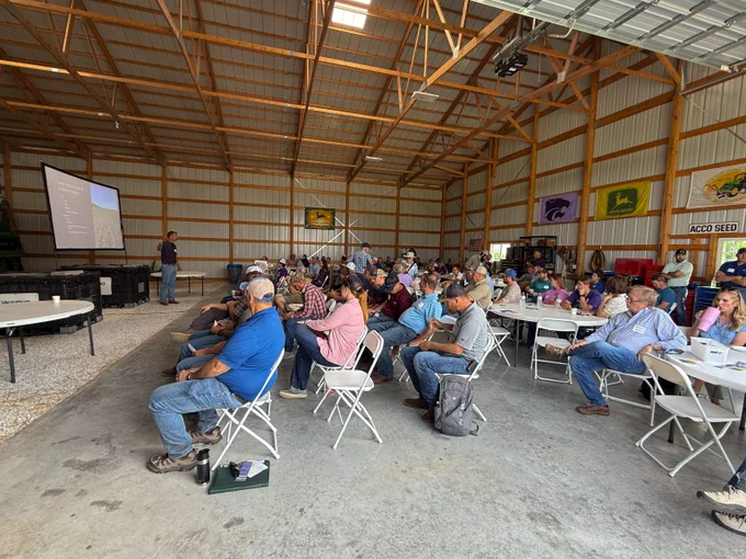 A group of people seated on folding chairs inside a large barn attending a presentation with a projector screen at the front.