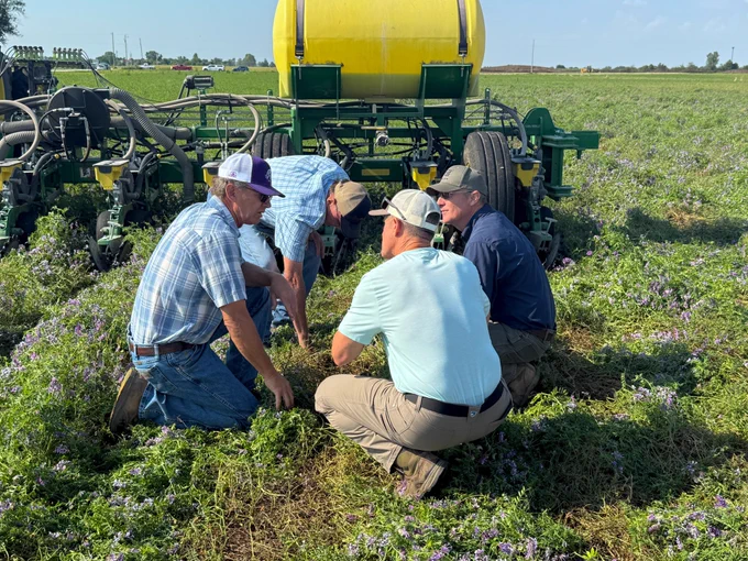Four men kneeling in a field examining soil and plants near agricultural equipment on a sunny day.