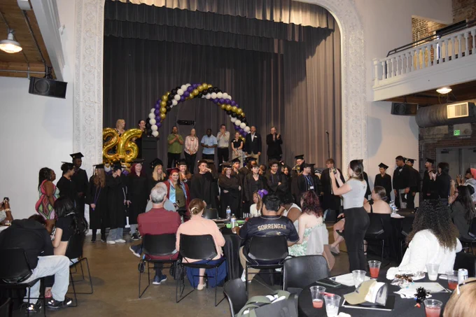 A large group of young people wearing caps and gowns stand in front of a stage decorated with a balloon arch in an auditorium. A seated crowd watches them.